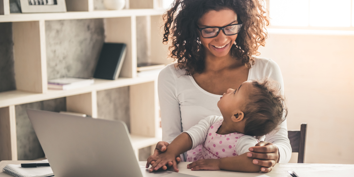 A mother holds her young child while working on a laptop at home as part of her guide to returning to work after maternity leave