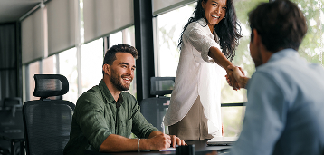 Two professionals shaking hands during a business meeting while a colleague smiles at the table